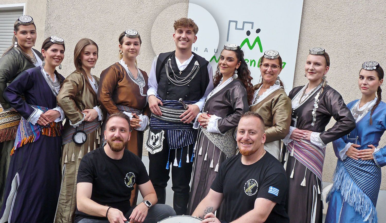 Eine Gruppe von Künstlern in traditionellen griechischen Trachten posiert mit zwei Männern in schwarzen T-Shirts vor einem Gebäude.