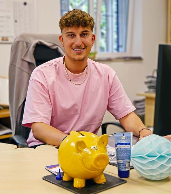 Eine Person in einem hellrosa T-Shirt sitzt an einem Schreibtisch mit einem gelben Sparschwein, einer Tube Lotion und einem blauen Papierornament.