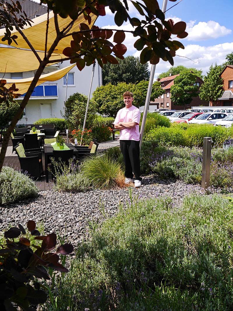 Eine Person in einem rosa Hemd und einer dunklen Hose steht mit verschränkten Armen auf einer sonnigen, landschaftlich gestalteten Terrasse.