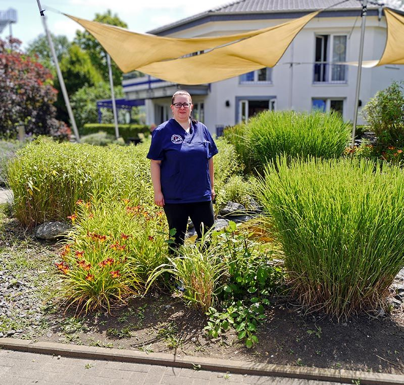Eine Person in blauer Uniform steht lächelnd inmitten üppiger grüner Pflanzen in einem Garten; im Hintergrund sind ein Gebäude und ein Sonnensegel zu sehen.