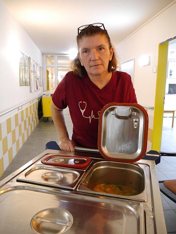 Ein Mitarbeiter in weinroter Uniform hält in einem sterilen, gefliesten Flur einen Speisenwärmer aus Metall auf einem Servierwagen offen.