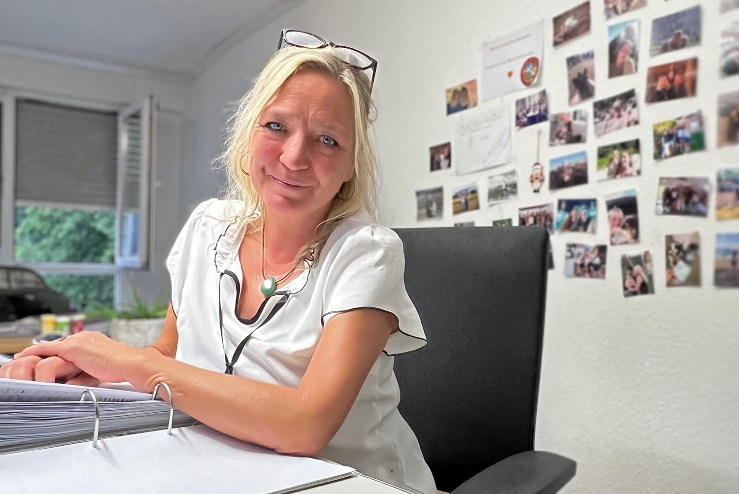 Eine Person mit blonden Haaren und Brille sitzt lächelnd an einem Schreibtisch; hinter ihr befindet sich eine Wand mit kleinen Fotografien.