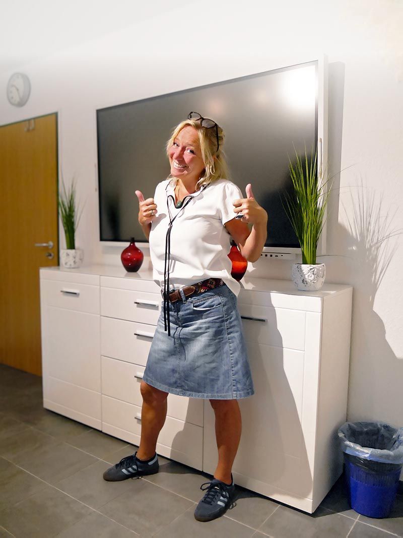 Eine Person in einem weißen Hemd und einem Jeansrock steht in einem Raum und zeigt mit beiden Händen den Daumen nach oben.