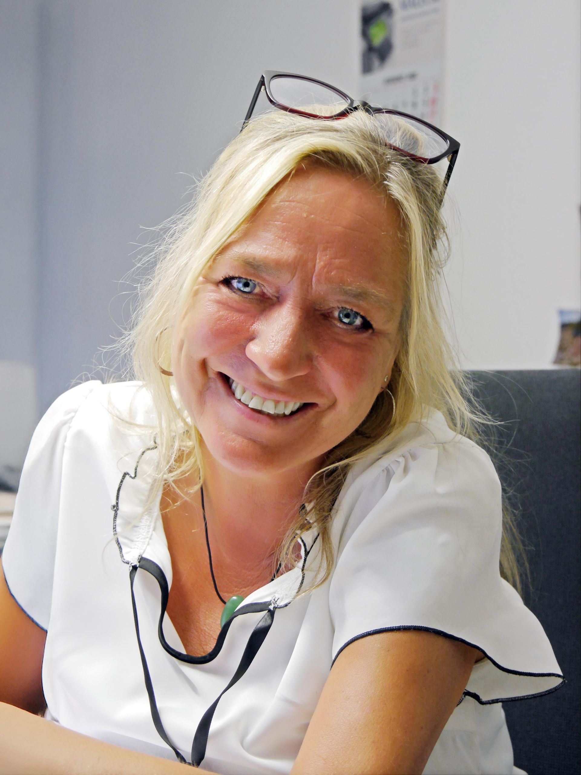 Eine lächelnde Person mit blonden Haaren und einer auf dem Kopf sitzenden Brille, die ein weißes Hemd mit dunklem Saum trägt.