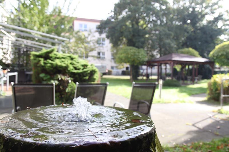 Im Vordergrund befindet sich ein kleiner Springbrunnen, im Hintergrund sind eine verschwommene Terrasse, Grünflächen und ein Pavillon zu sehen.