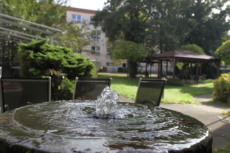 Im Vordergrund plätschert ein kleiner Springbrunnen, dahinter befinden sich Terrassenstühle und ein Pavillon in einem üppigen, grünen Garten.