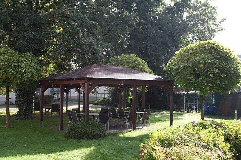 In einem grünen Garten, umgeben von gepflegten Bäumen, steht ein hölzerner Pavillon, darunter sind Tische und Stühle aufgestellt.