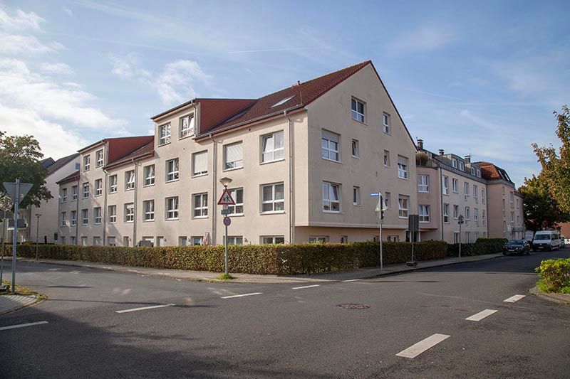 An einer Straßenecke steht an einem sonnigen Tag ein hellfarbiges, mehrstöckiges Wohnhaus mit rotem Ziegeldach.