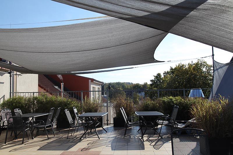 Eine Dachterrasse mit Metalltischen und -stühlen unter einem großen grauen Sonnenschutz vor hellem, sonnigem Hintergrund.