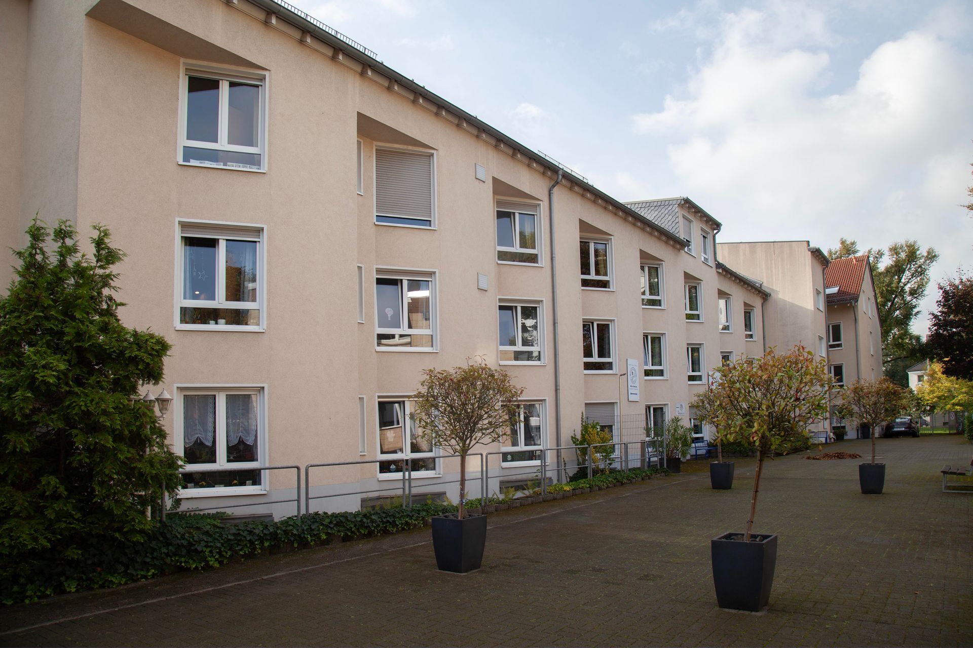 Ein mehrstöckiges beigefarbenes Gebäude mit Reihen rechteckiger Fenster, umgeben von einem gepflasterten Innenhof mit Topfbäumen.
