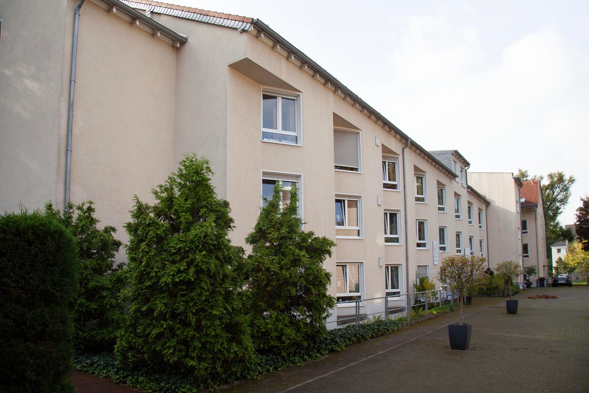 Ein hellfarbiges, dreistöckiges Wohnhaus mit mehreren Fenstern, umgeben von Bäumen und einem gepflasterten Innenhof.