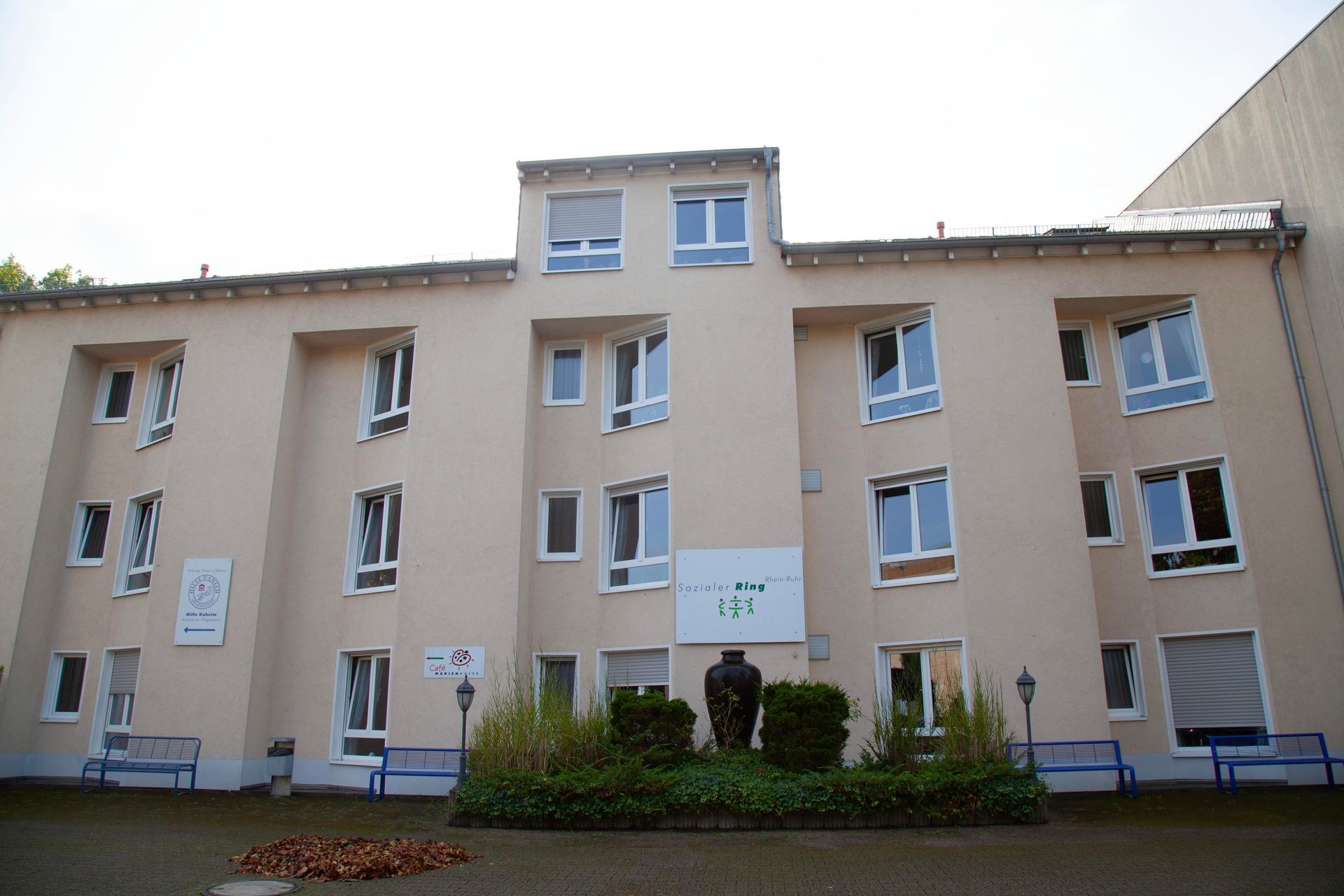Ein vierstöckiges, hellfarbiges Gebäude mit mehreren Fenstern, einem zentralen Eingangsbereich und Bänken im Erdgeschoss.
