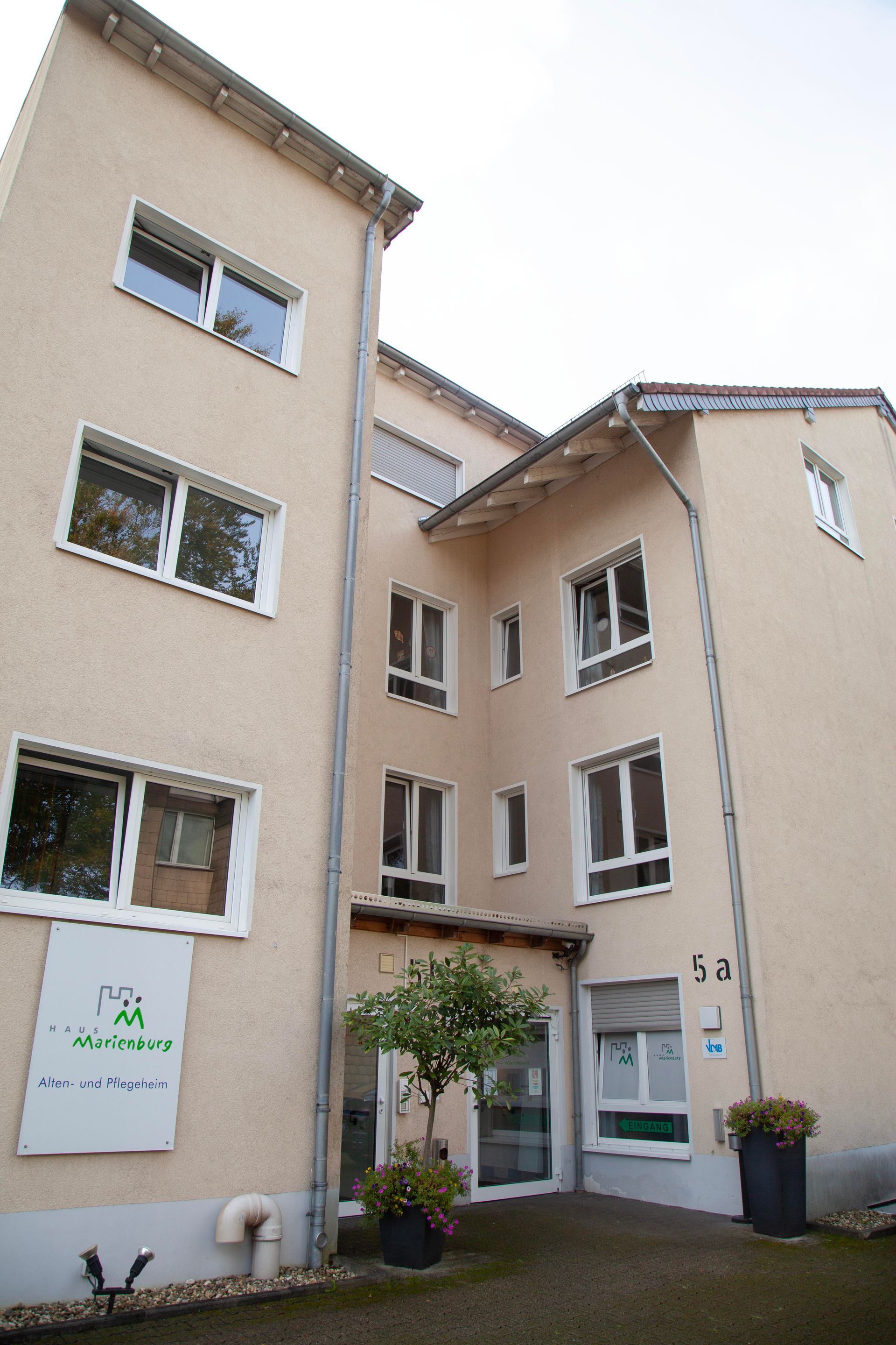 Eine Ansicht aus der Froschperspektive auf ein beigefarbenes, mehrstöckiges Gebäude mit weiß gerahmten Fenstern, einem zentralen Eingang und zwei Topfpflanzen.