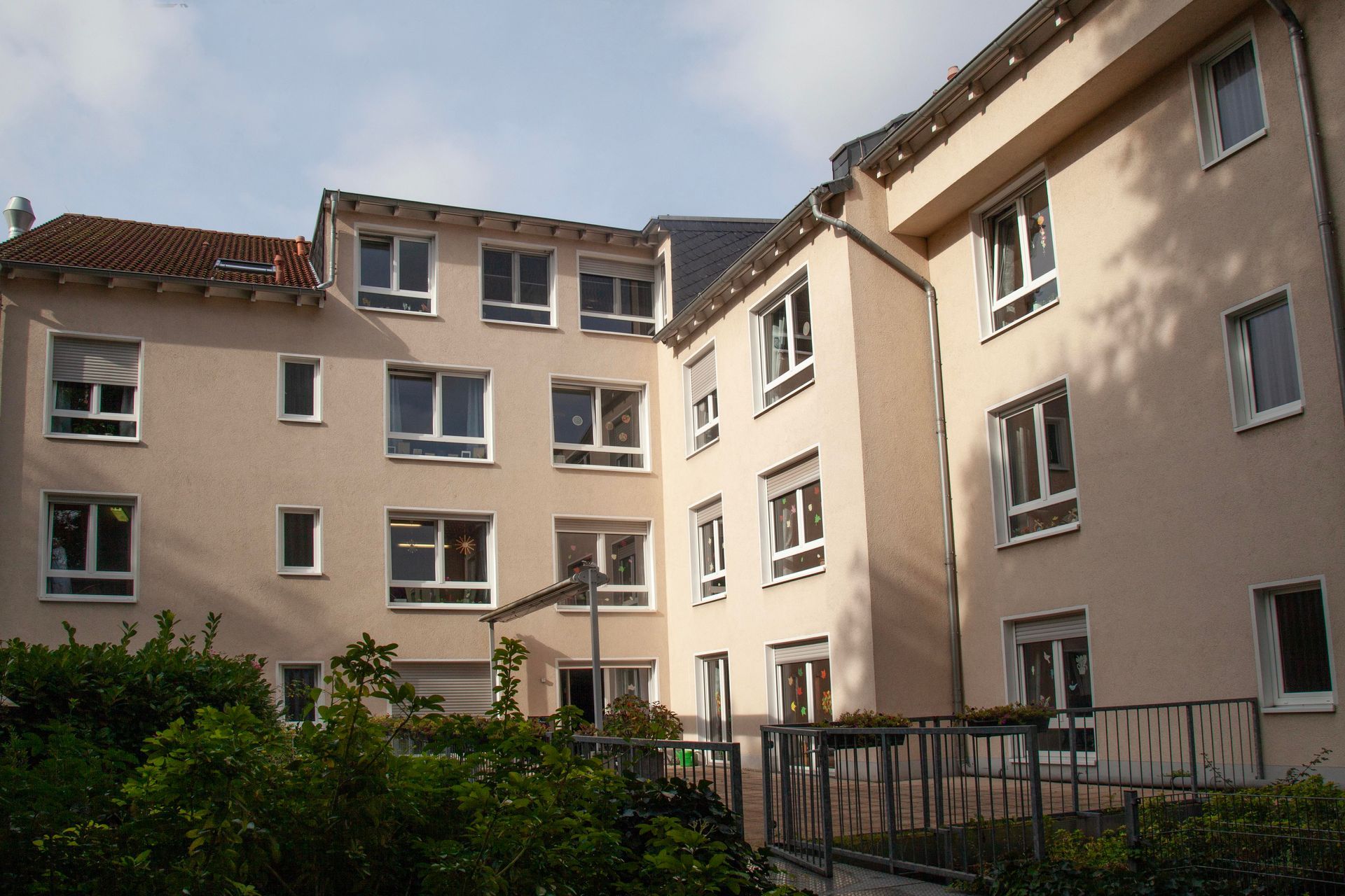 Ein mehrstöckiges, beigefarbenes Wohnhaus mit einem zentralen Innenhof und Grünflächen im Vordergrund unter bewölktem Himmel.
