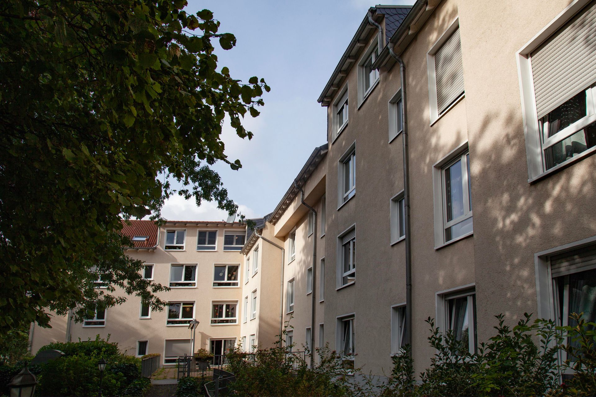 Ein mehrstöckiges beigefarbenes Wohnhaus mit weißgerahmten Fenstern, das an einem sonnigen Tag teilweise von grünen Bäumen beschattet wird.