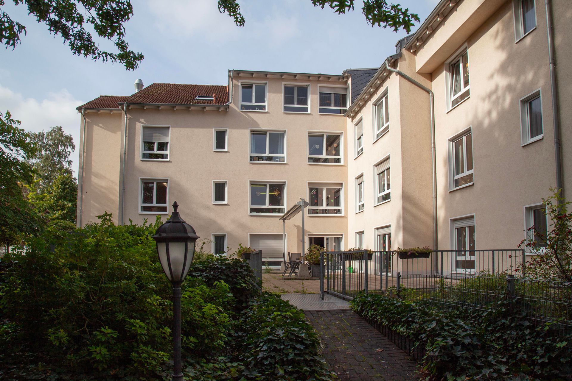 Ein cremefarbenes, mehrstöckiges Wohnhaus mit Garten, Laternenpfahl und Grünpflanzen im Vordergrund.