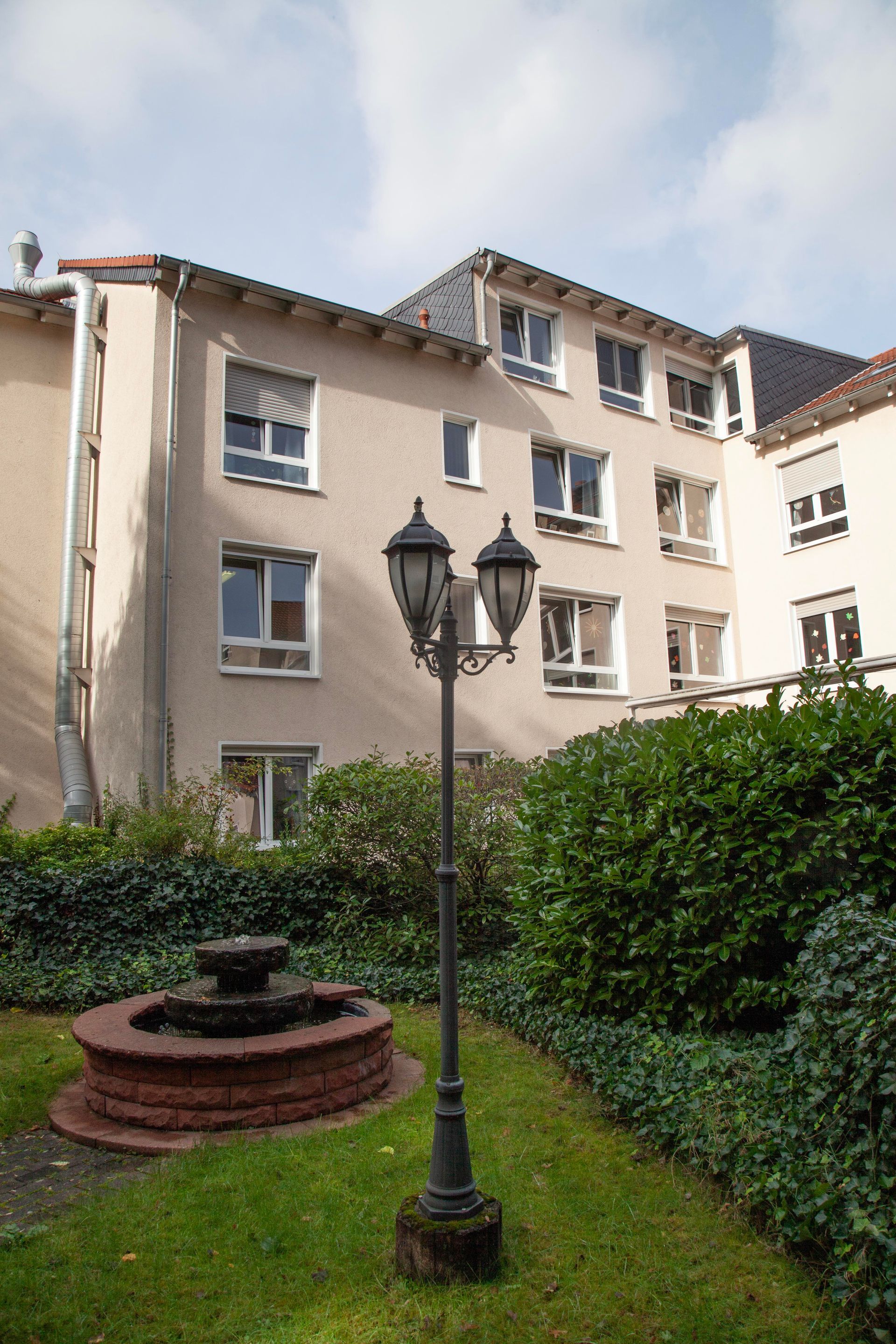 In einem grünen Garten vor einem dreistöckigen, hellen Wohnhaus steht unter bewölktem Himmel ein kleiner Steinbrunnen.