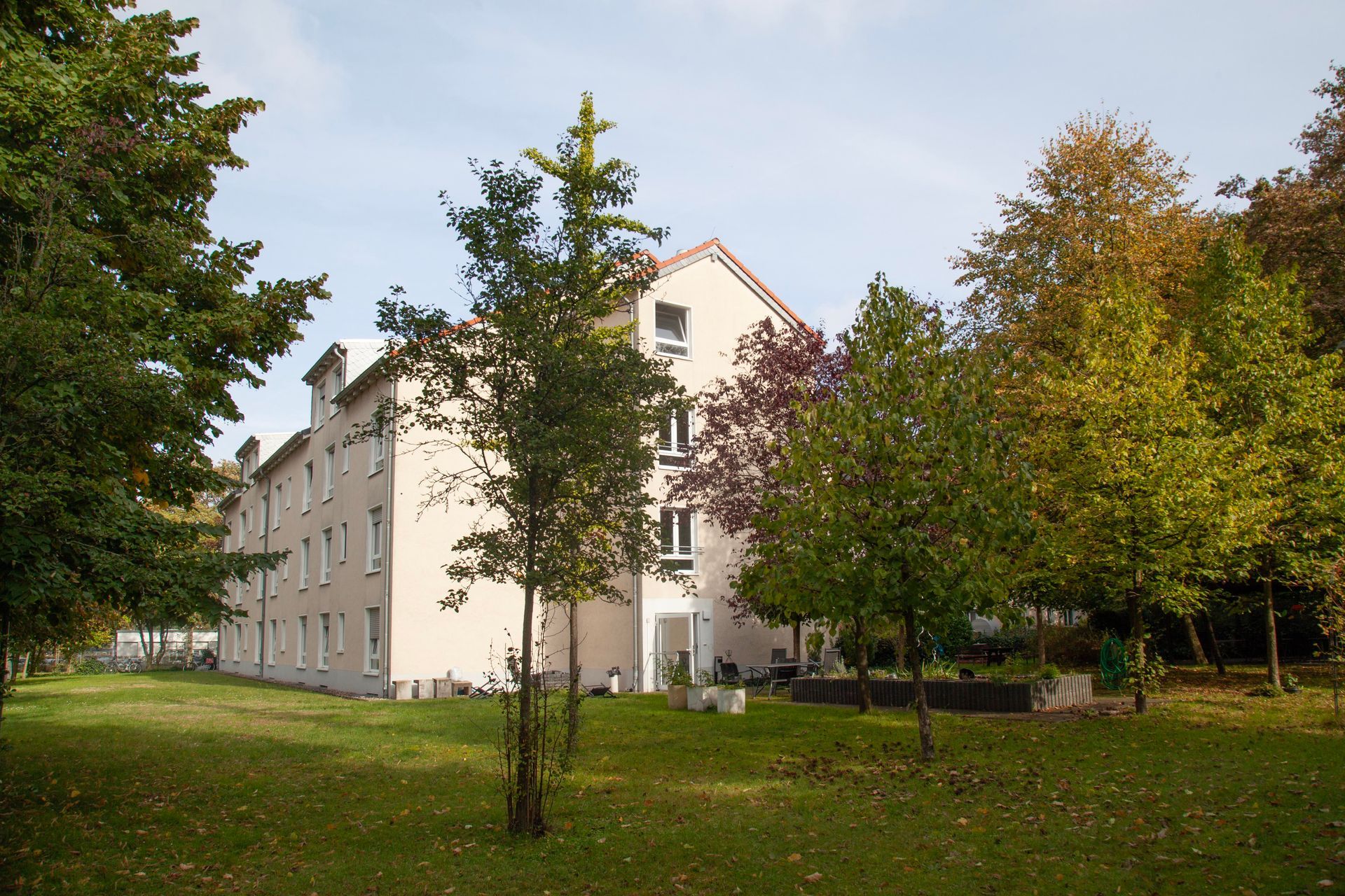In einer grasbewachsenen Fläche, umgeben von verschiedenen grünen und gelblichen Bäumen, steht ein mehrstöckiges, hellrosa Gebäude.