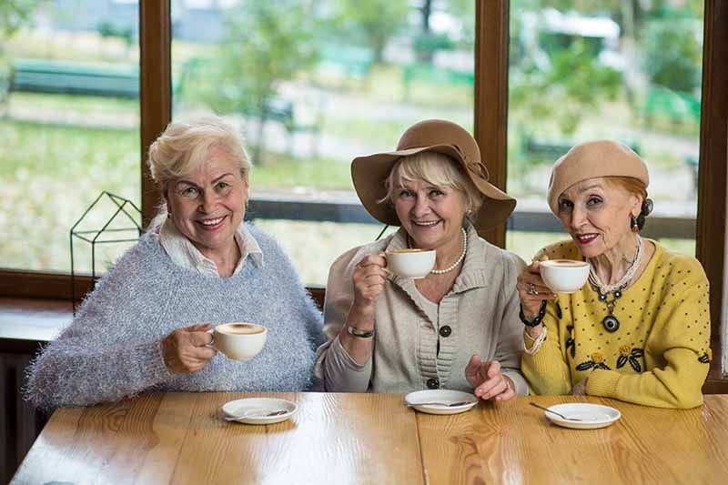 Drei Seniorinnen trinken Kaffee und lächeln dabei keck in die Kamera, Stockfoto
