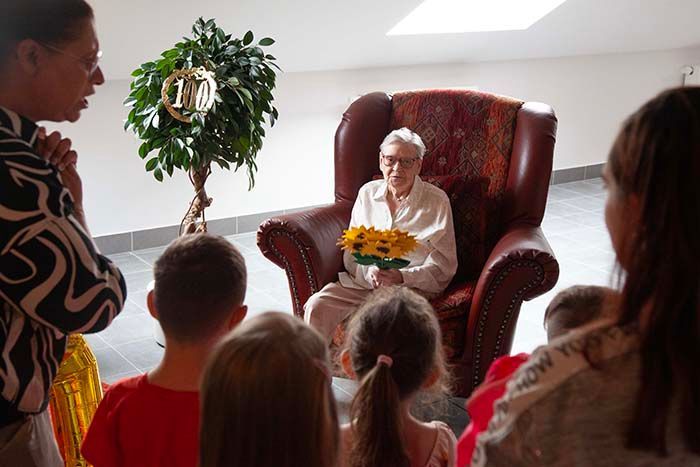 Eine Person sitzt in einem Ledersessel und hält Sonnenblumen in der Hand, umgeben von einer Gruppe von Menschen in einem hellen, festlichen Raum.