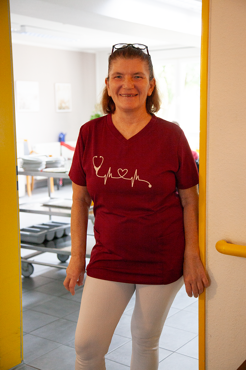 Eine lächelnde Person in einem weinroten T-Shirt mit Stethoskop- und Herzmotiv steht im Türrahmen eines Raumes mit Metallwagen.