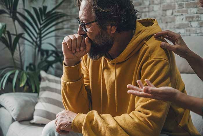 Mann im mittleren Alter sitzt verzweifelt auf einer Couch. Eine Person neben ihm, von der nur die Hände zu sehen sind, will ihm Mut zusprechen.