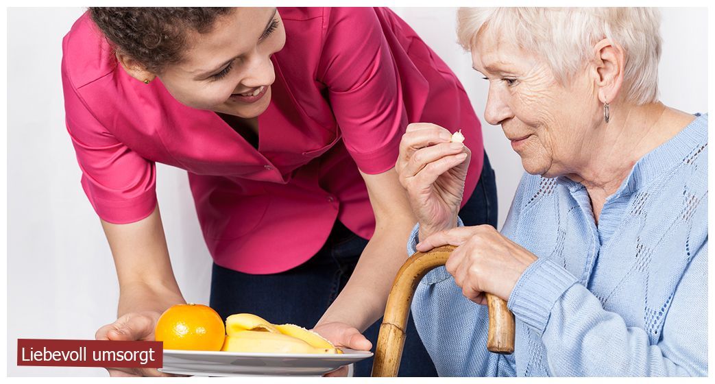 Eine Pflegekraft in rosa Uniform hält einen Teller mit Obst und bietet einer Person, die einen Gehstock benutzt, eine Pille an.