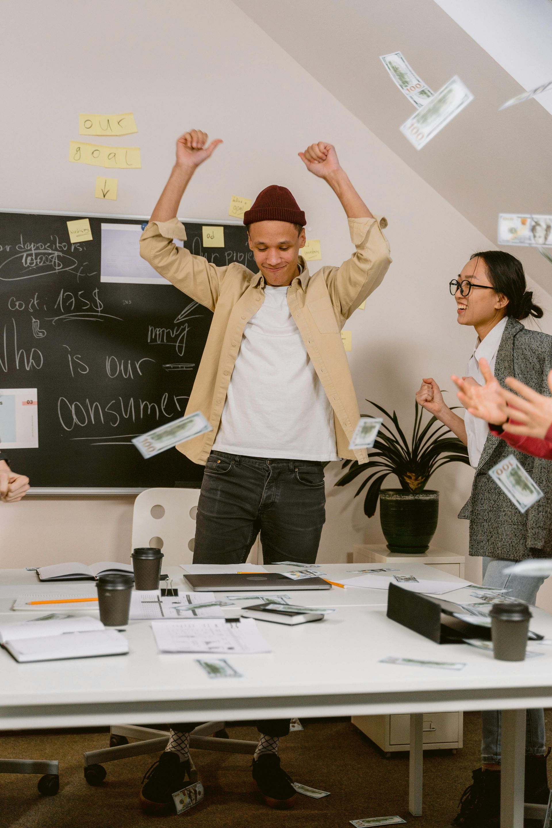 People celebrating with money falling in an office. A man in a beanie and jacket raises arms, others clap.
