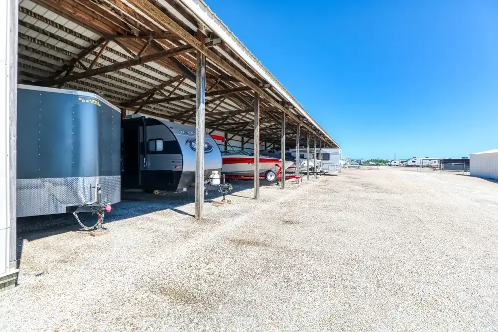 Covered storage units with gravel ground, housing trailers and a boat under a blue sky.