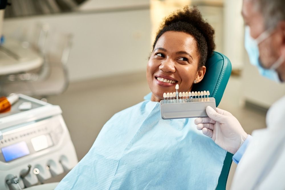 A woman is sitting in a dental chair while a dentist holds a tooth color chart. — Burleigh Heads Denture Clinic Gold Coast in Burleigh Heads, QLD