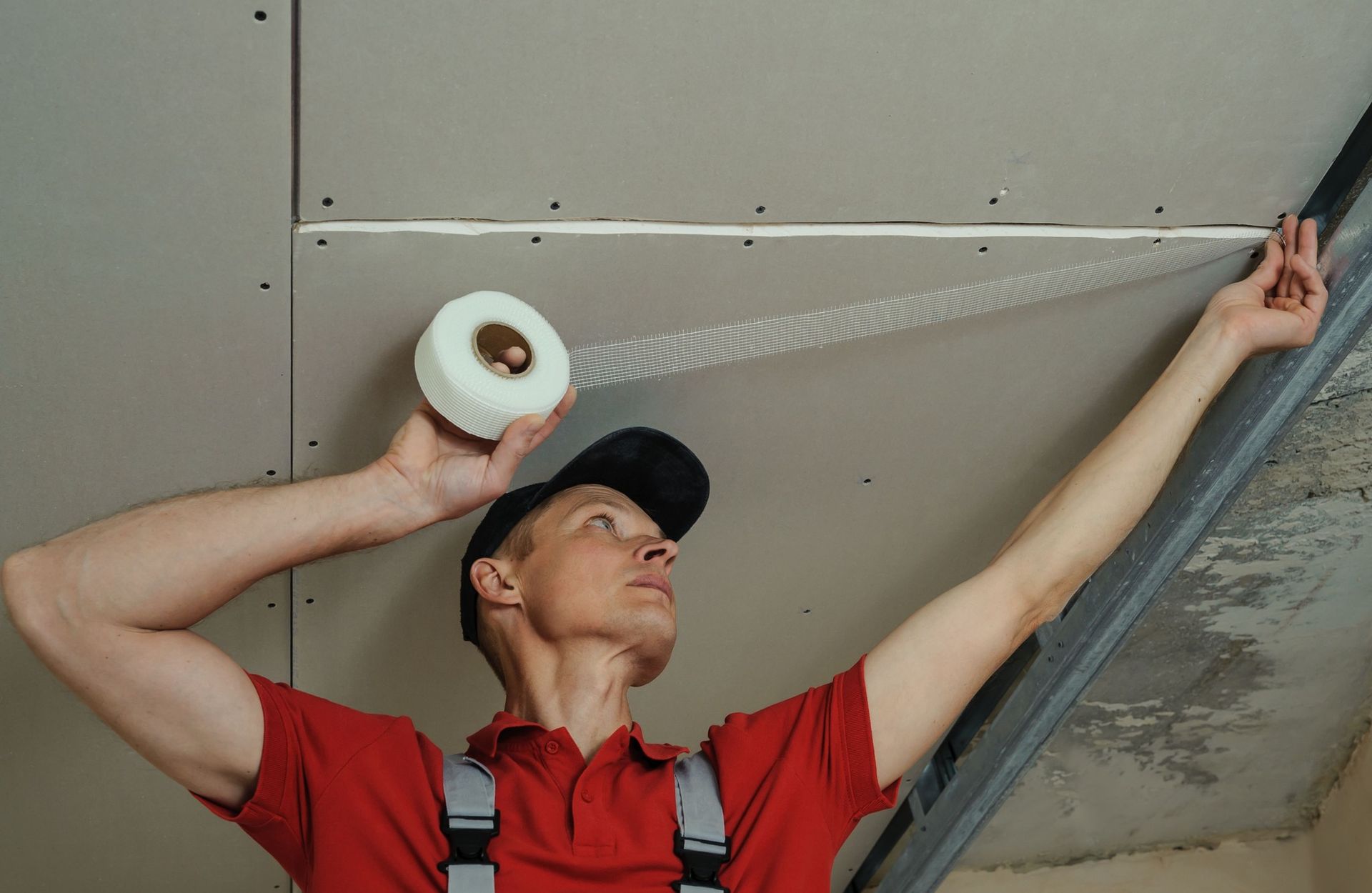 A worker in a red shirt applies adhesive mesh tape to a drywall seam on a ceiling.