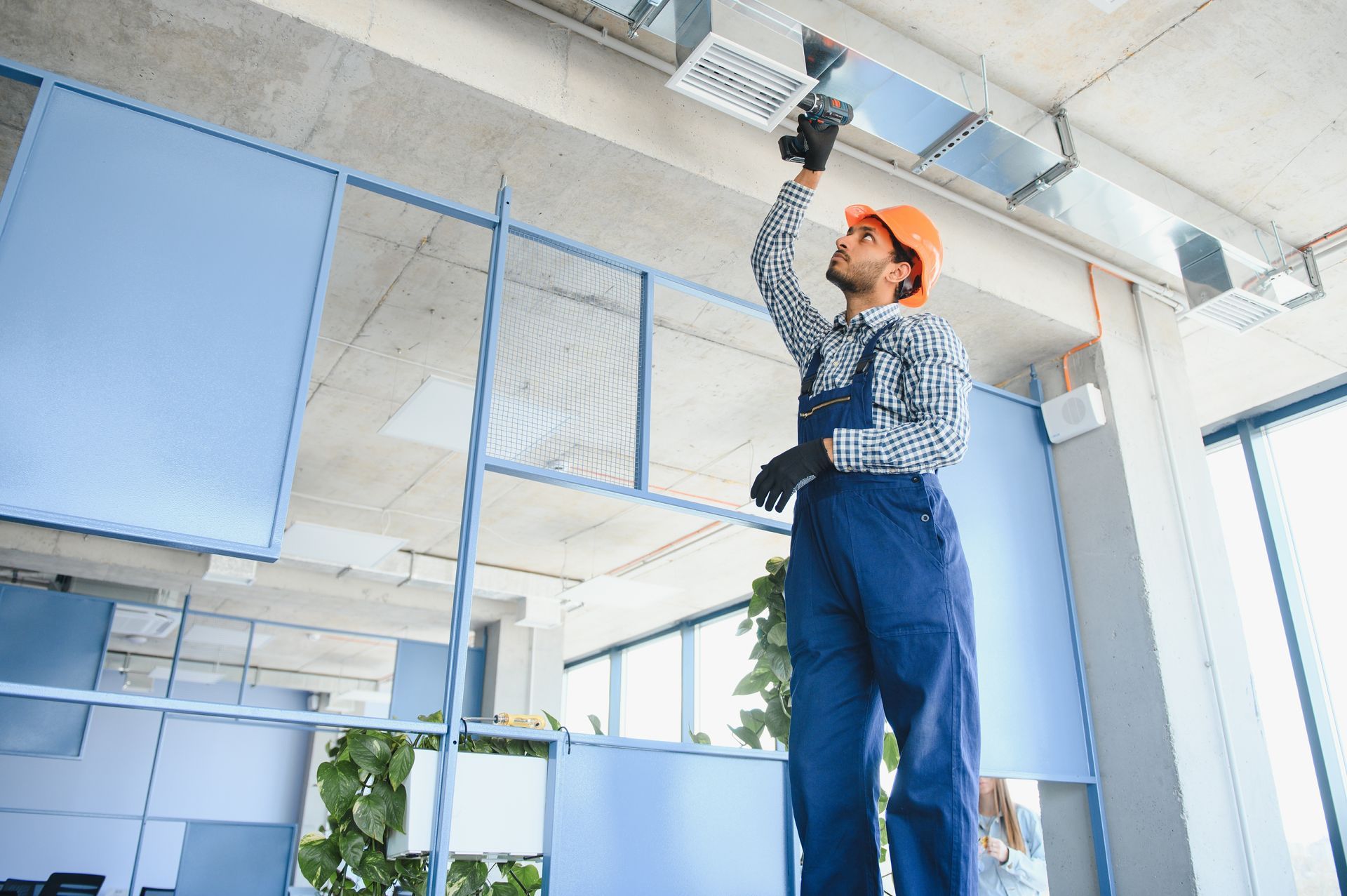 Construction worker in blue coveralls and orange hard hat installs HVAC ductwork on ceiling.