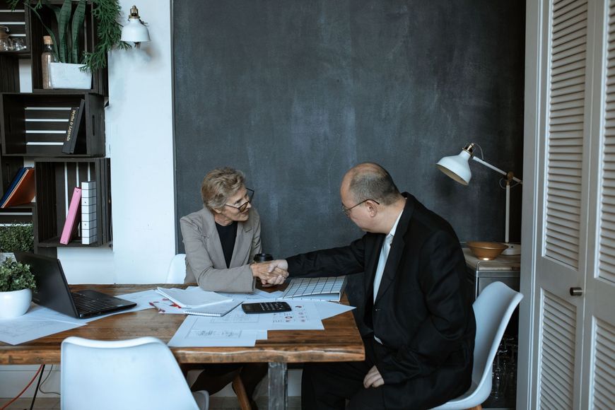 Two people in an office discussing papers at a table, with a laptop and desk lamp nearby.