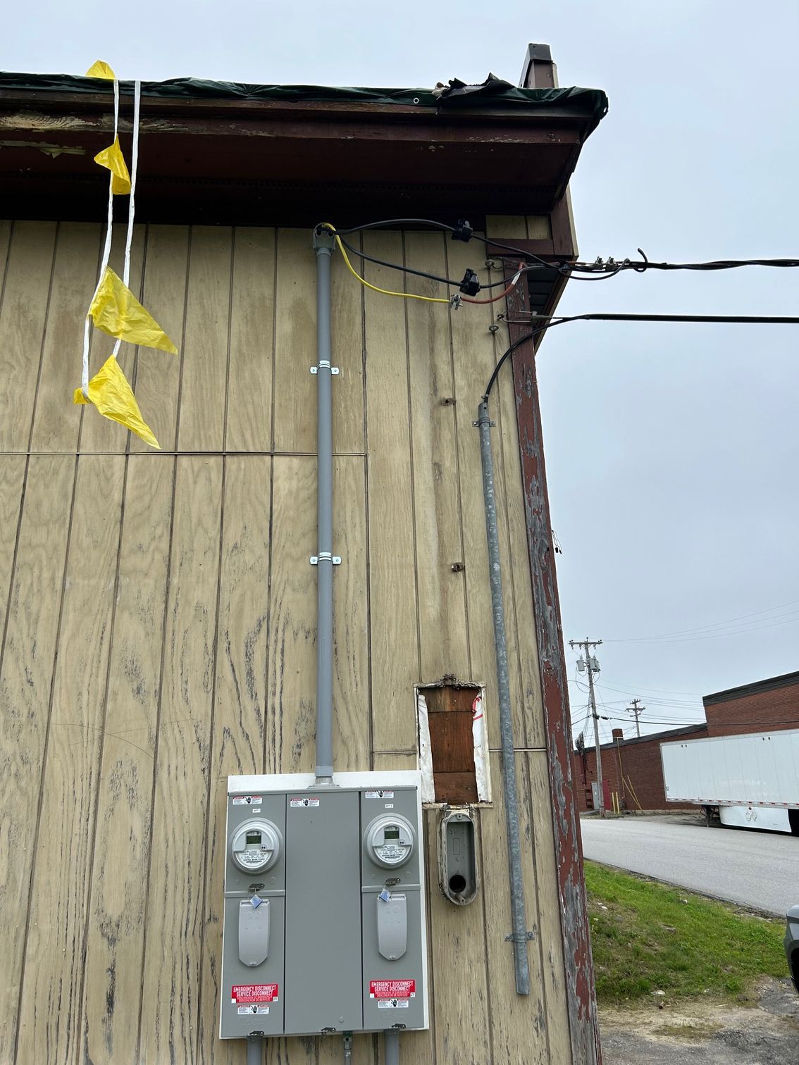 Exterior of weathered building with utility meters, conduit pipes, and yellow caution tape near the roofline