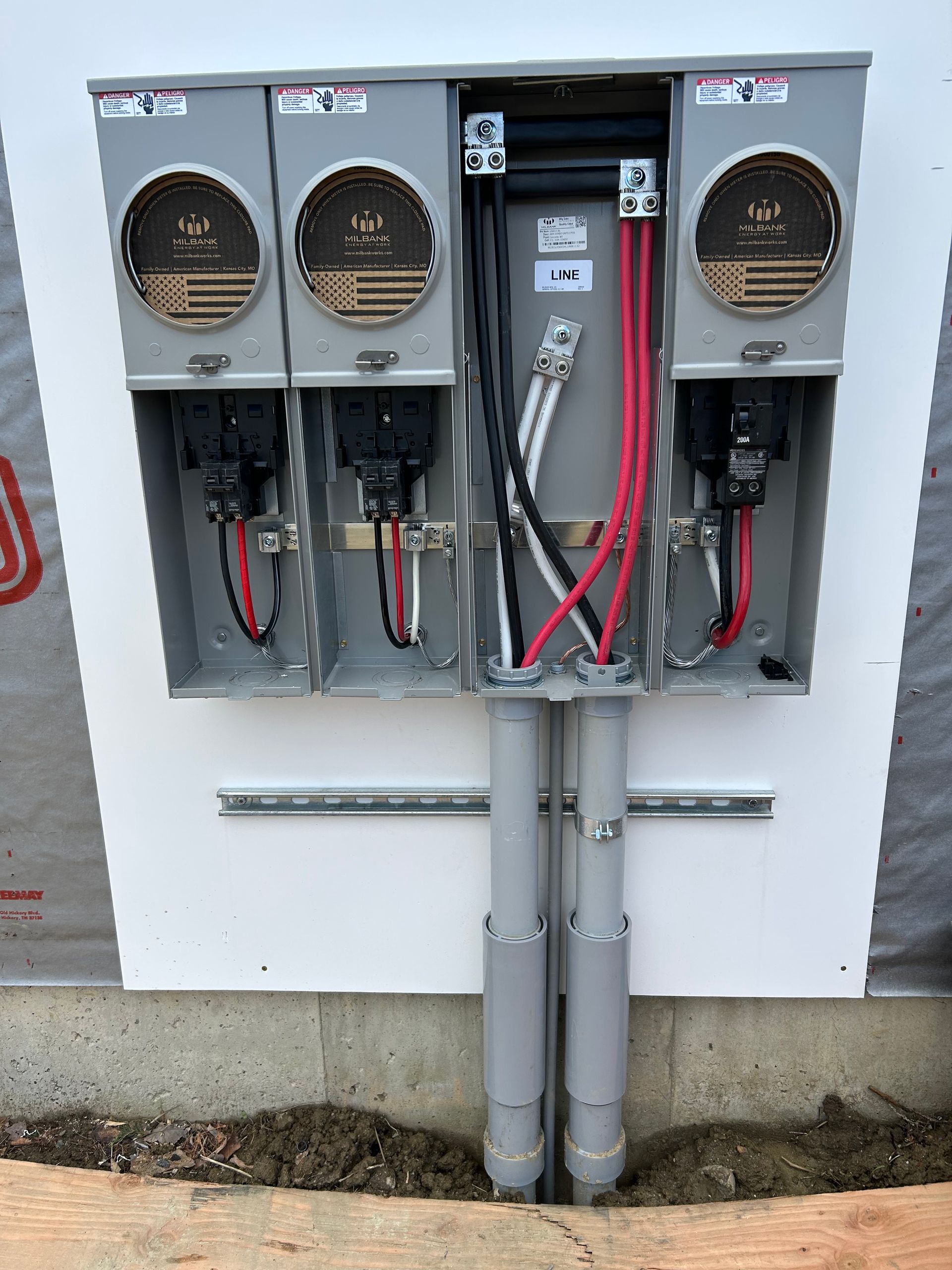 Outdoor electrical utility meter and service panel boxes mounted on a white wall with exposed conduit and wiring.