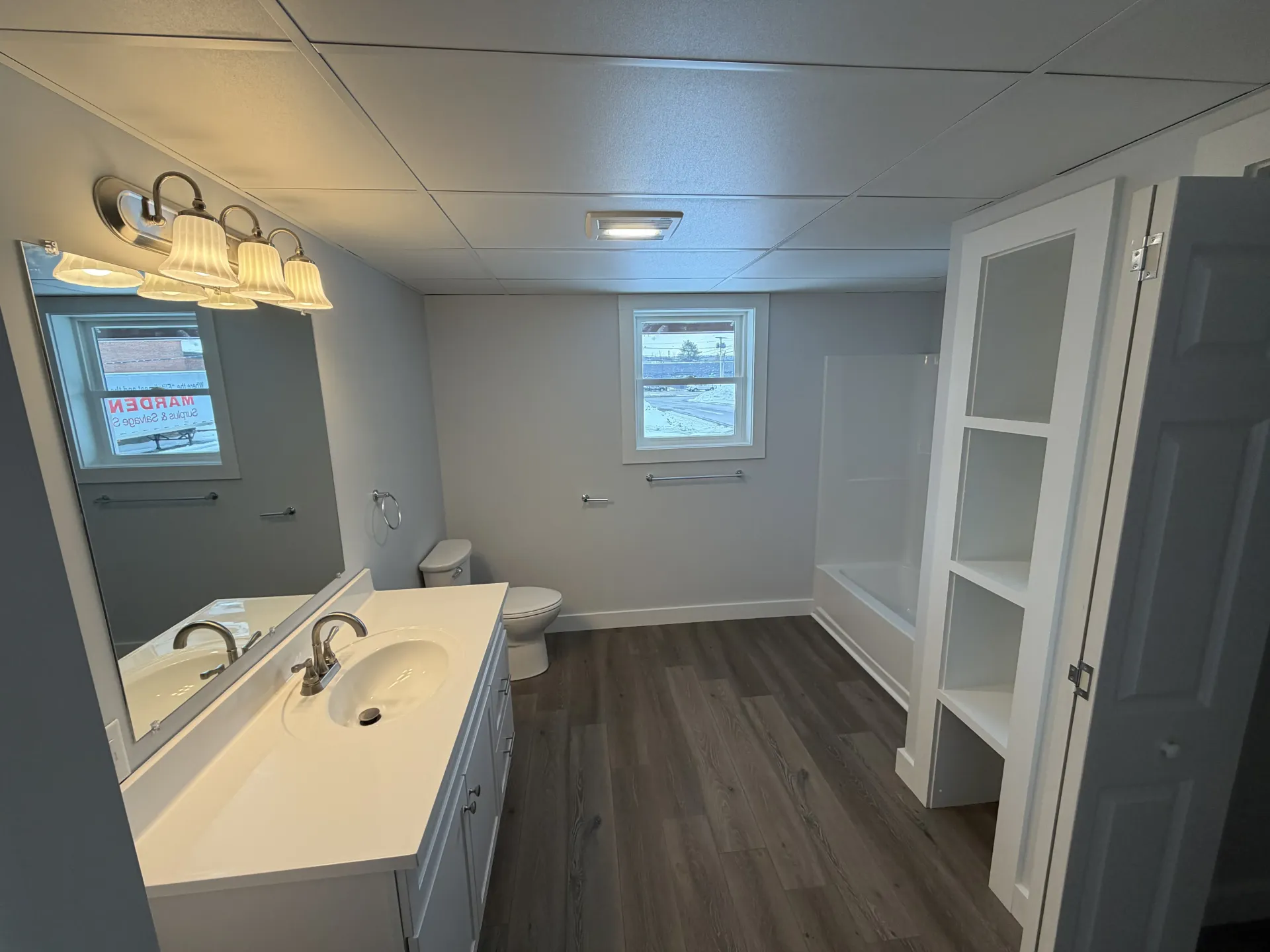 Empty bathroom interior with white vanity, dark wood floor, large mirror, and built-in shelving.