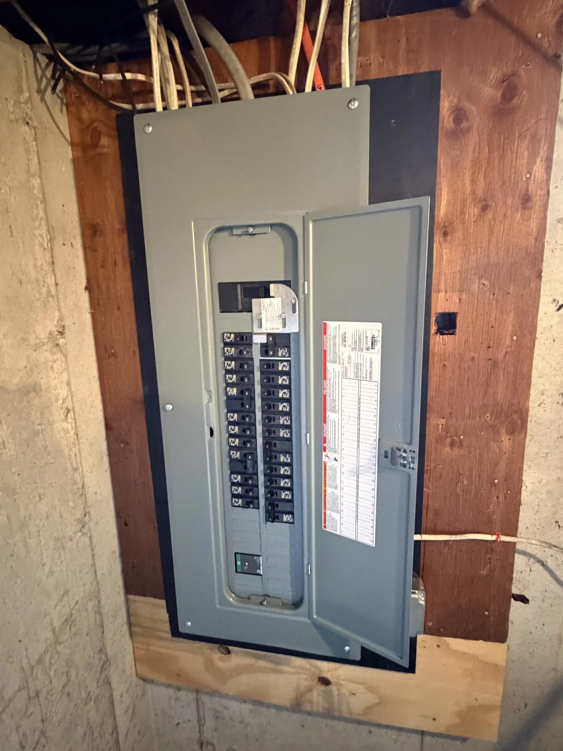 Open electrical breaker panel in unfinished wall, with door ajar and exposed wiring above.