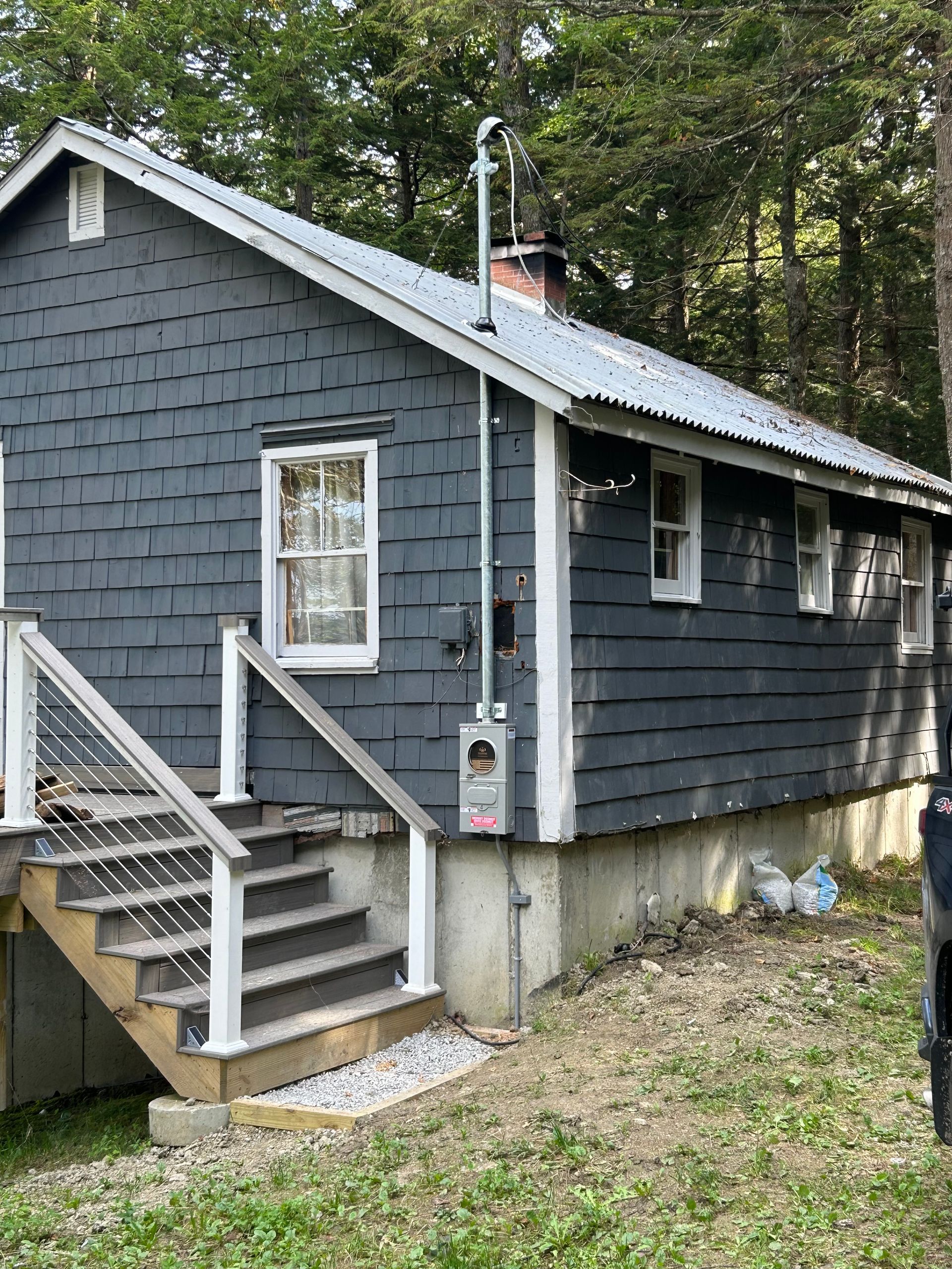 Small gray-shingled house with white trim, front stairs, and a wooded yard.