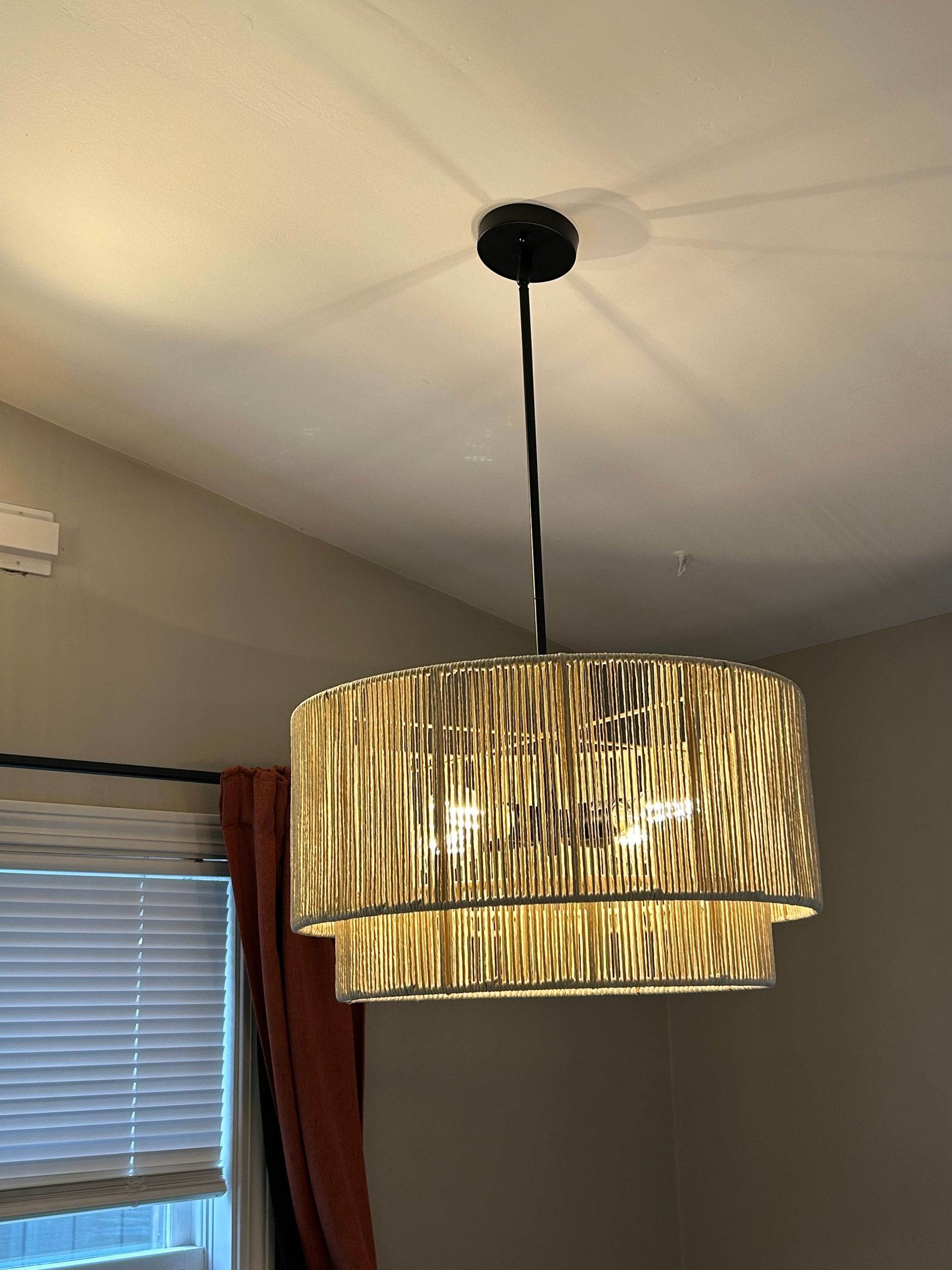 Gold woven drum pendant light hanging from the ceiling above a window with blinds and red curtains