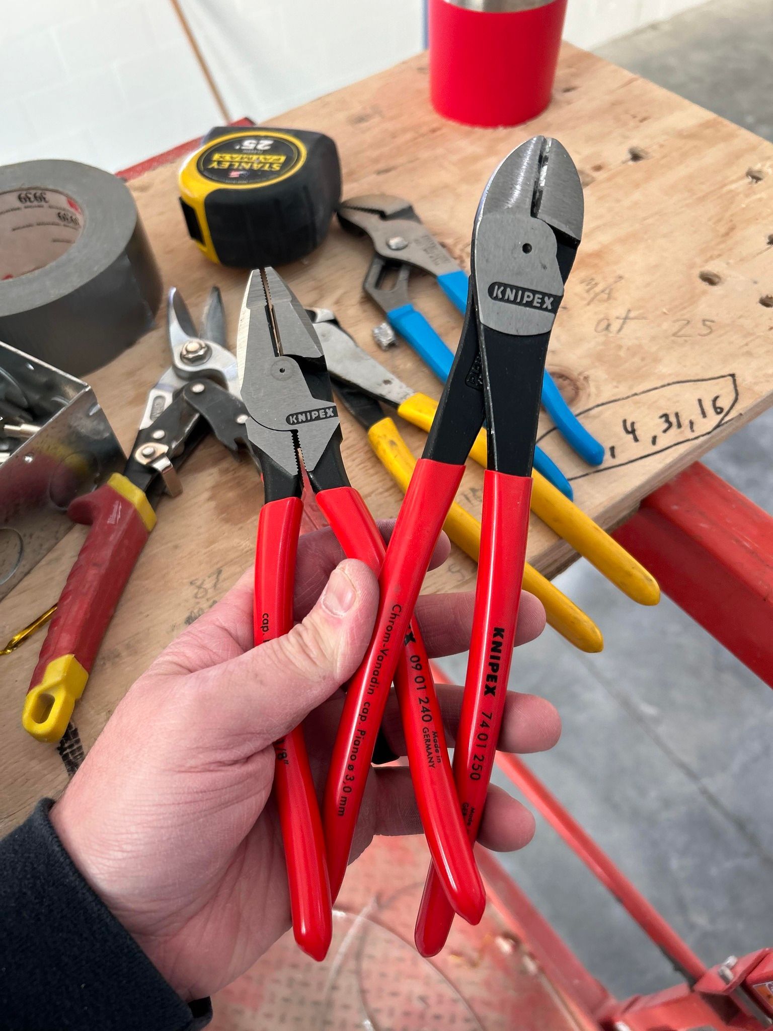 Hand holding red-handled pliers over a workbench with assorted tools and a red cup in the background