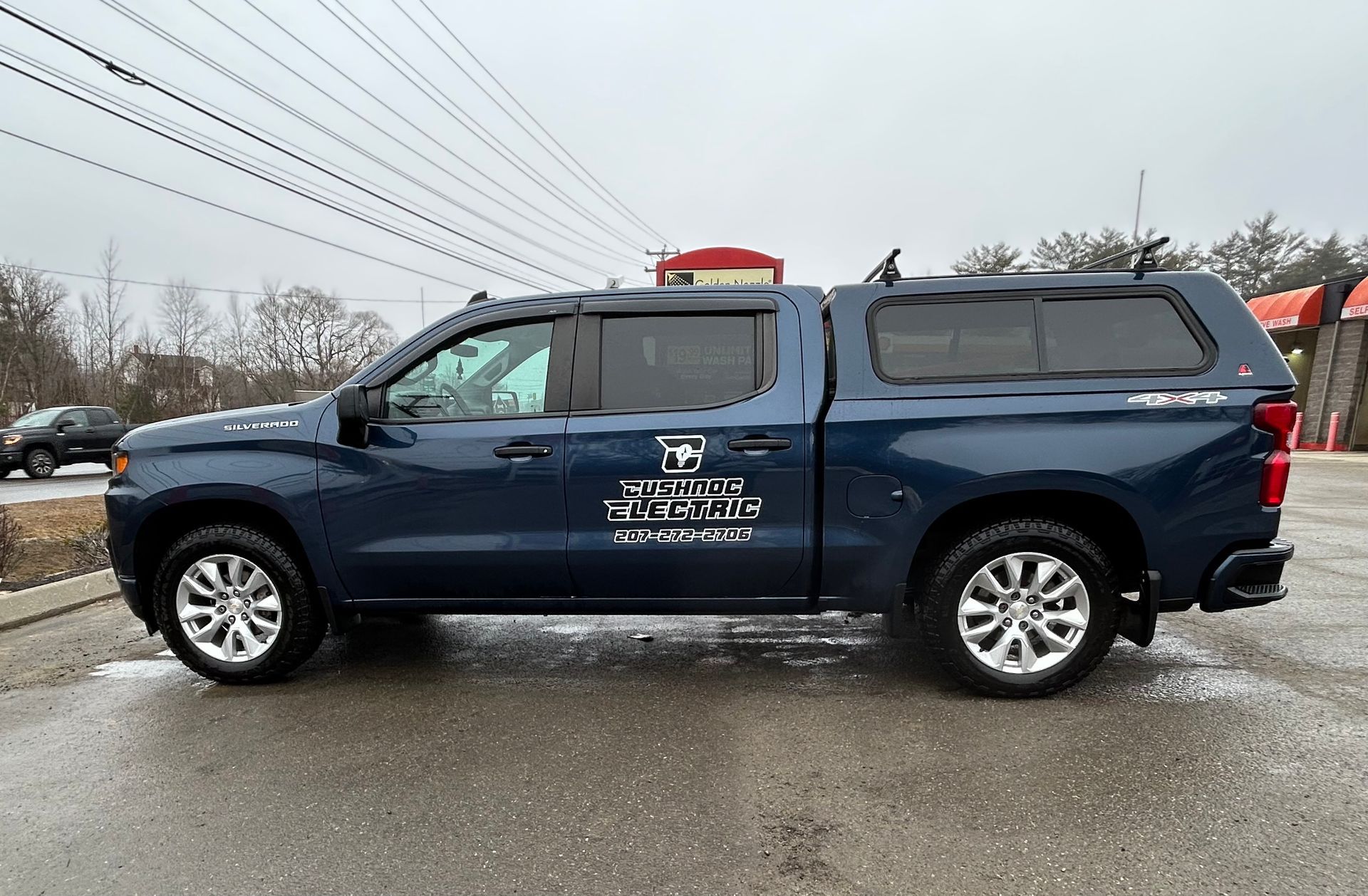 Blue pickup truck with a camper shell parked on a wet lot under an overcast sky