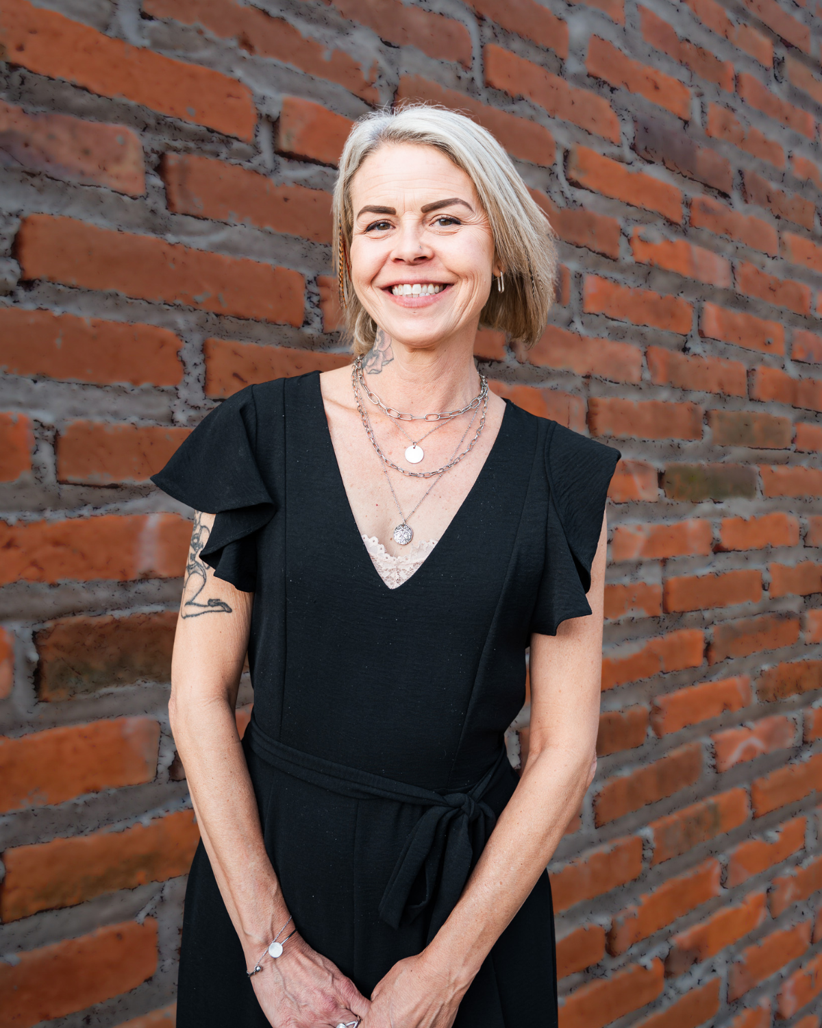 Smiling person in a black dress standing against a red brick wall.