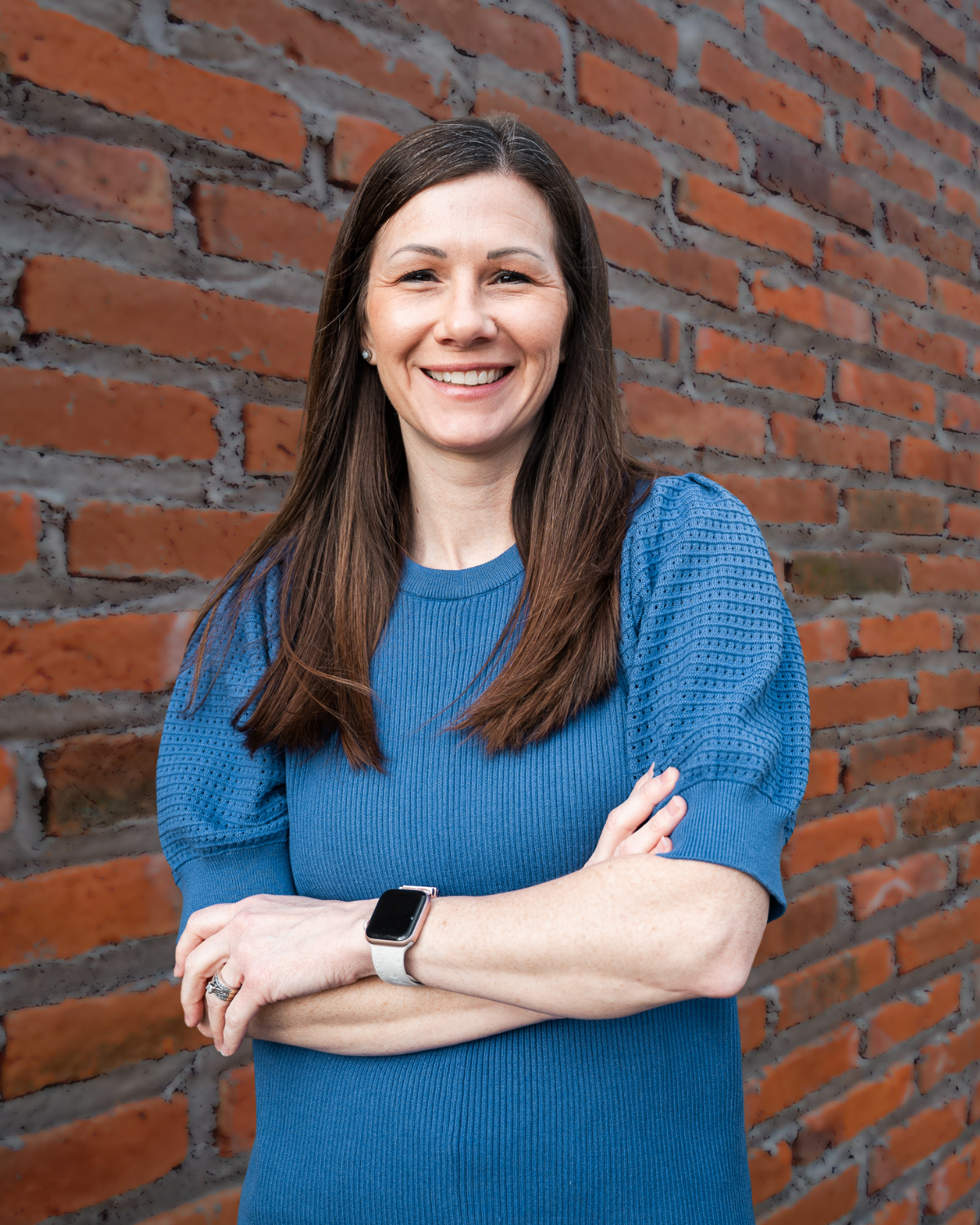 Smiling person in a blue sweater with crossed arms standing against a brick wall