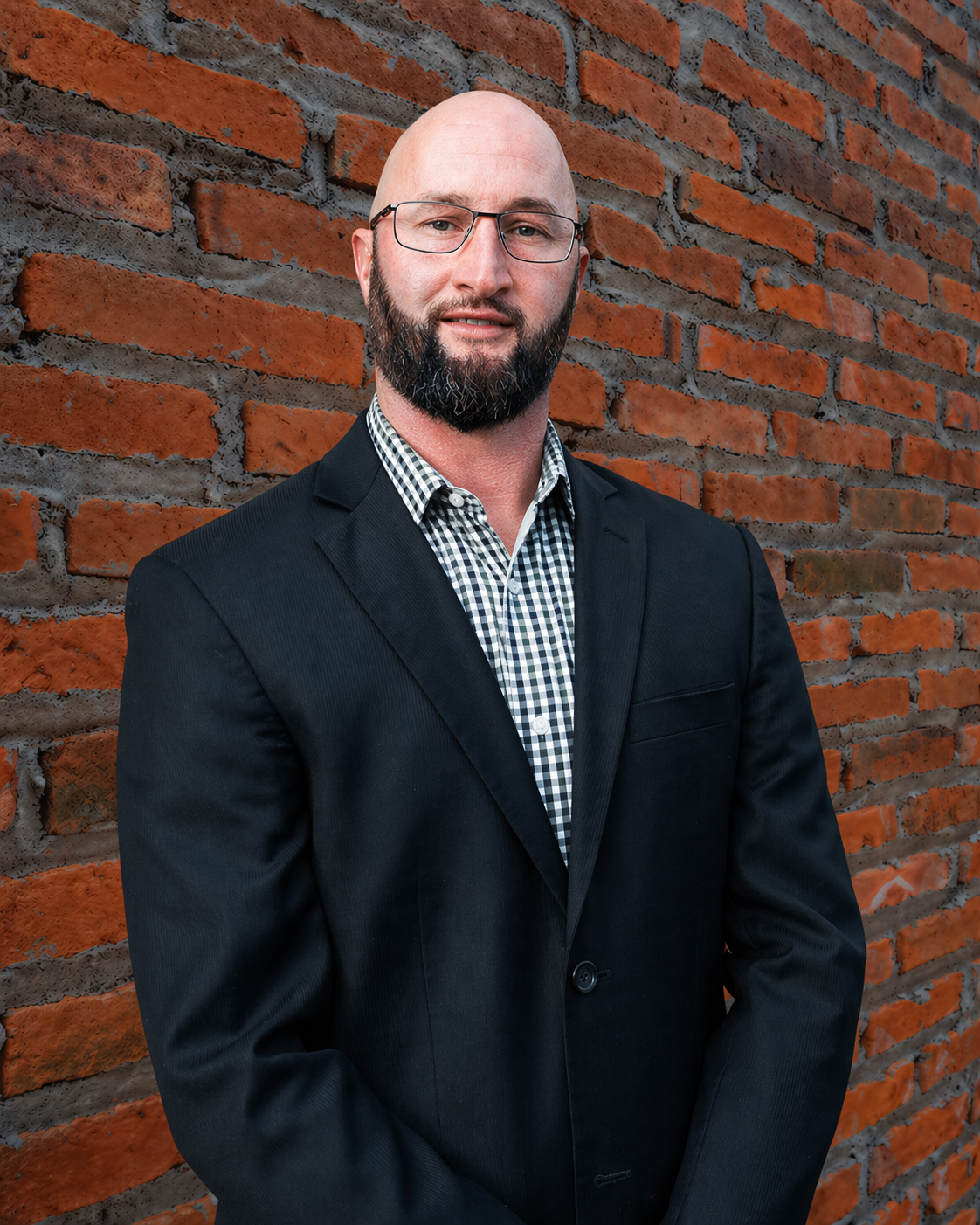 Man in a dark suit standing against a brick wall, wearing glasses and a checkered shirt.