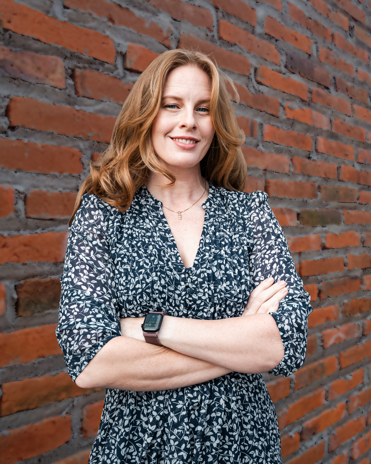 Woman with crossed arms smiling beside a red brick wall, wearing a dark patterned dress.