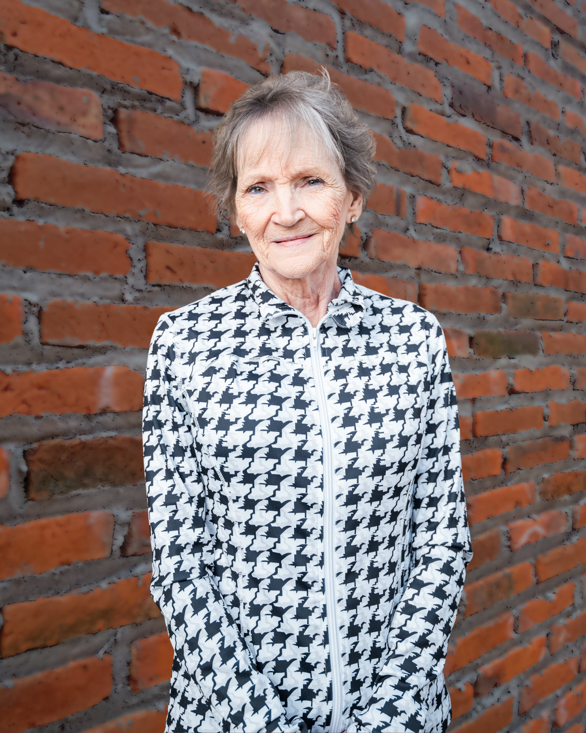 Smiling person in a black-and-white patterned shirt standing against a brick wall