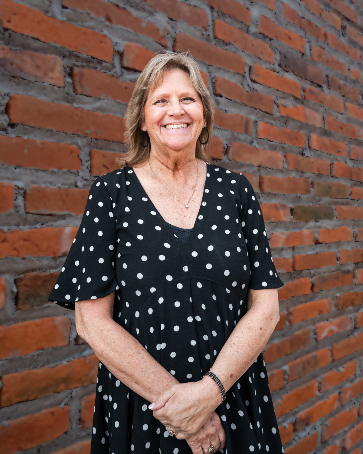 Smiling woman in a black polka dot dress standing against a brick wall.