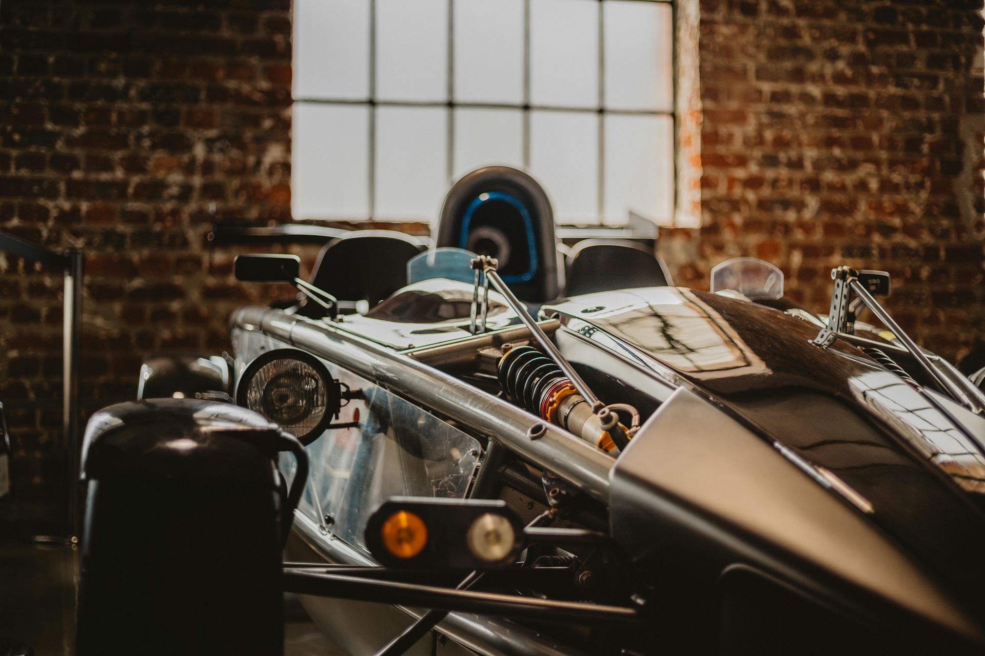 Silver and black open-wheel sports car with visible suspension, parked in front of a brick wall and window.