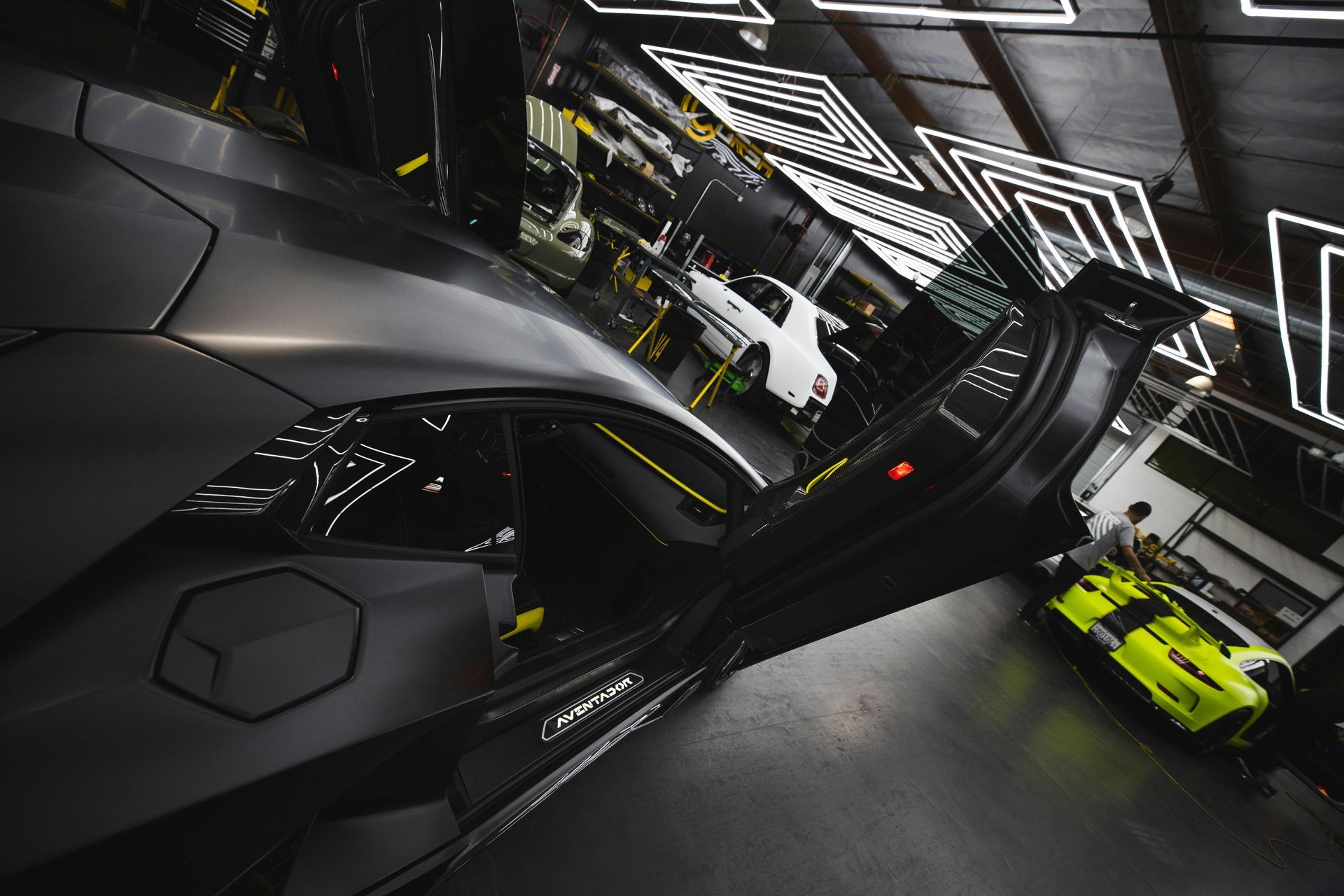 Gray sports car with open door in a garage, a neon yellow car, and other vehicles in the background.