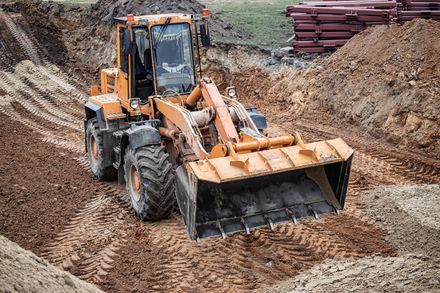 Orange front loader digging dirt in a construction site with piles of soil and stacked materials in the background